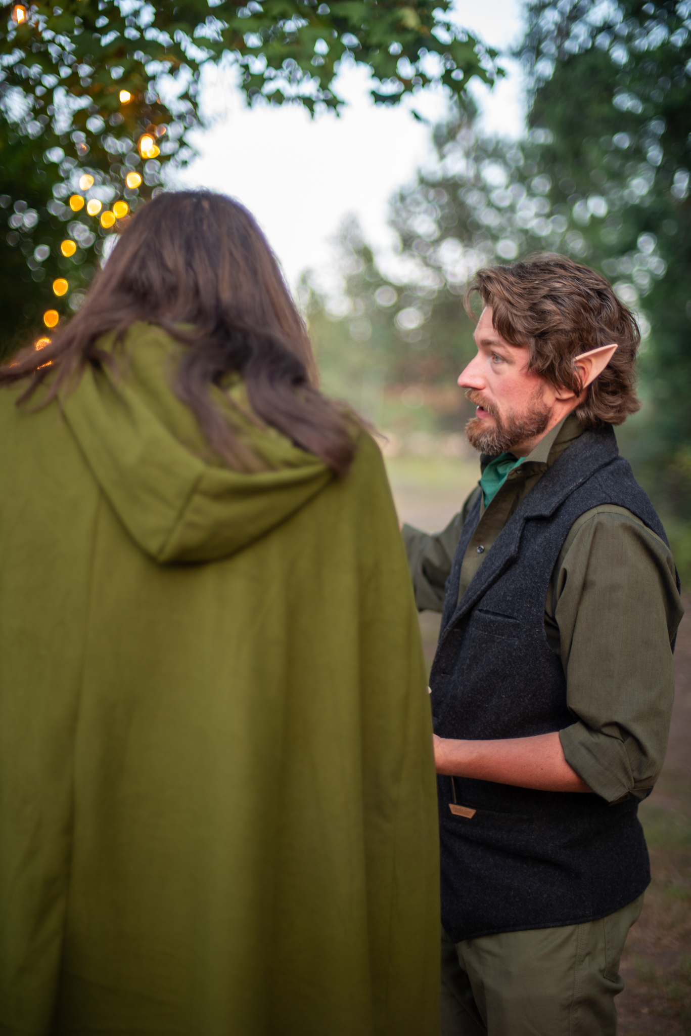 A guest in elf ears and a wool waistcoat talking with a friend in a green hooded cloak