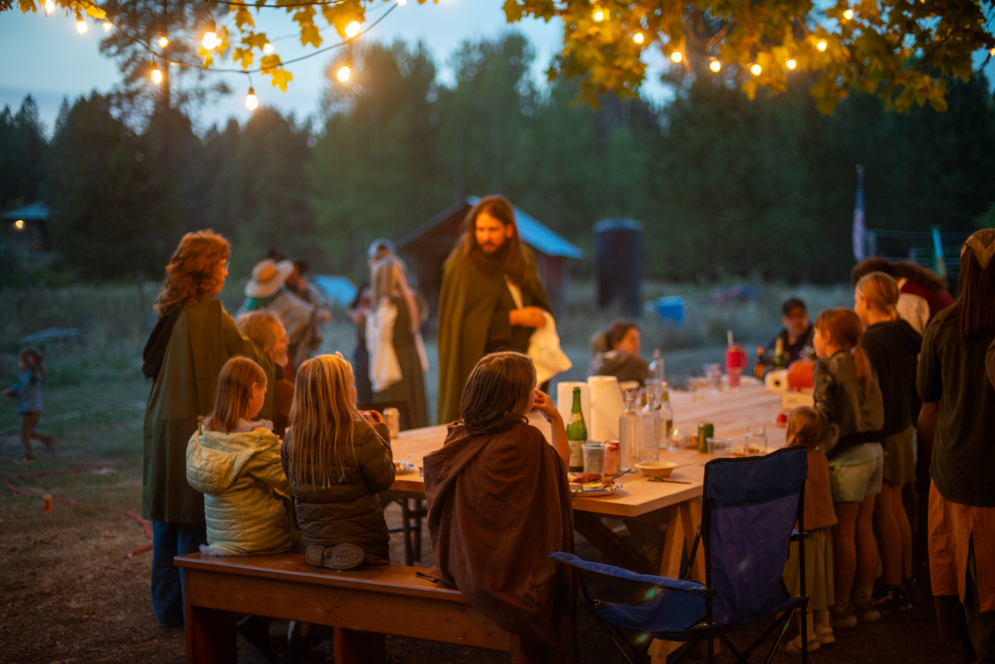 Guests gathered around a long wooden table beneath the party tree at dusk, strung with warm lights