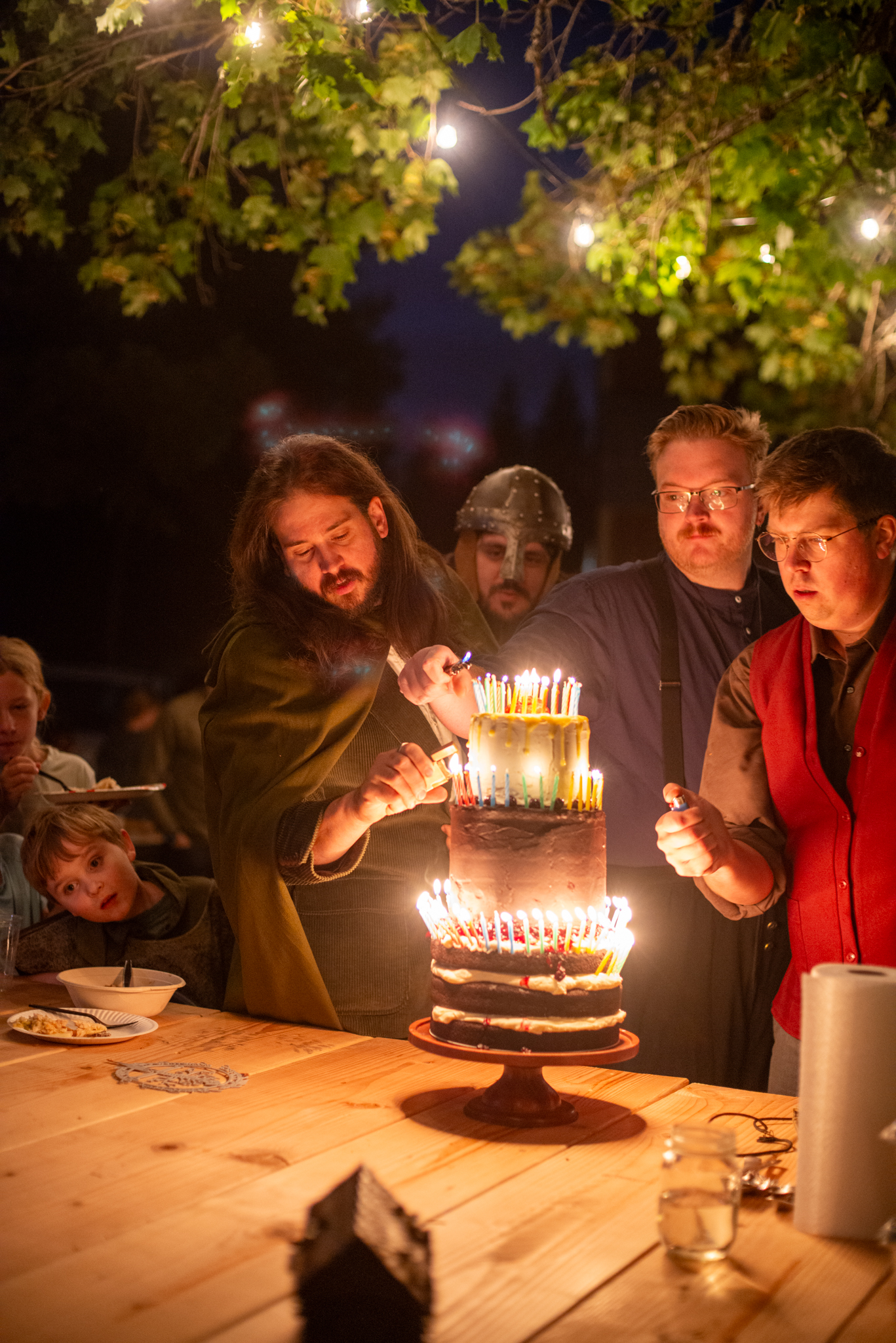 Guests lighting eleventy-one candles on a tiered birthday cake after dark